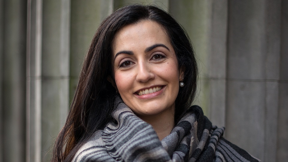 A woman with long dark hair and a scarf smiles warmly at the camera.